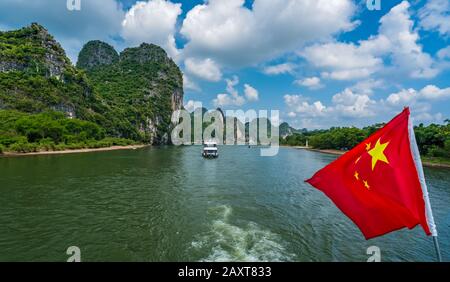 Rote chinesische Nationalflaggen flattern auf einem Mast auf dem Deck des Sightseeing-Boatts voller Touristen, die auf einer Reise auf dem herrlichen Fluss Li von starten Stockfoto