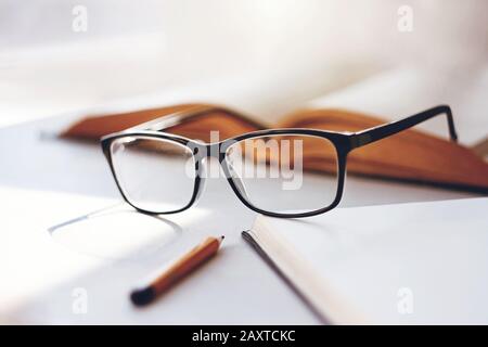 Auf einem weißen Tisch liegt ein offenes Buch, eine elegante, schwarz umrandete Brille, ein Notizbuch für Notizen und ein Bleistift, das vom Licht der Mittagssonne beleuchtet wird. Stockfoto