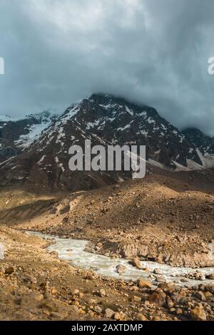 Der Fluss Chandra, der vor einem Berg mit Sonnenlicht fließt, der durch die Wolken zieht. Stockfoto