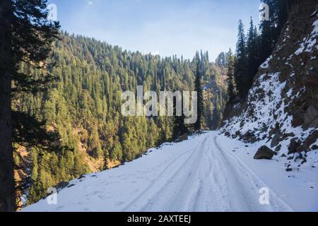 Eine schneebedeckte Straße mit Reifenprofilen auf der dunklen Seite des Berges. Stockfoto