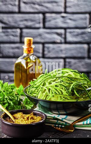 Spirogierte Zucchini Spaghetti, Zoodles in einer schwarzen Schüssel auf einem grauen Betontisch mit Sauce Pesto und Zutaten mit einer Ziegelwand im Hintergrund, Stockfoto