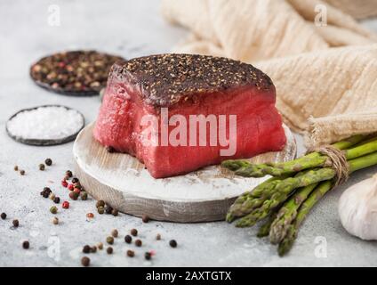 Scheibe Roher Rinderoberseite mit Salz und Pfeffer auf runder Hackbrett mit Spargelspitzen und Knoblauch auf hellem Küchentisch. Stockfoto