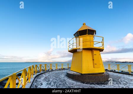 Kleiner gelber Leuchtturm an der Küste von Reykjavik, Island. Das Meer, der blaue Himmel und die schneebedeckten Berge sind im Hintergrund zu sehen. Stockfoto