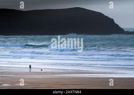 Eine Fernsicht auf einen einsamen Hundespender und ihren Hund, der an einem eisigen kalten Tag am Fistral Beach in Newquay in Cornwall am Ufer spaziert. Stockfoto