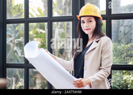 Porträt der schönen asiatischen Frau Architektenbauer mit gelbem Helm Harthut Studie Plan der Räume, ernsthafte Bauingenieurarbeiten w Stockfoto