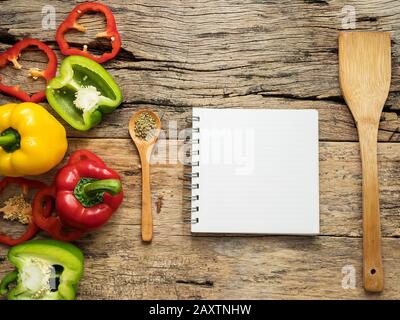 Flach von leeren Rezept kochen Buch und Utensilien mit Kräutern und bunten Paprika über Holz- Hintergrund. Ansicht von oben mit der Kopie Raum. Essen Rezept ein Stockfoto