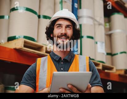 Erfolgreicher Lagerleiter lächelt und arbeitet während der Aufnahme eines digitalen Tabletts, um den Bestand und die Bestände mit weißem Helm und Sicherheitsjacke zu überprüfen Stockfoto