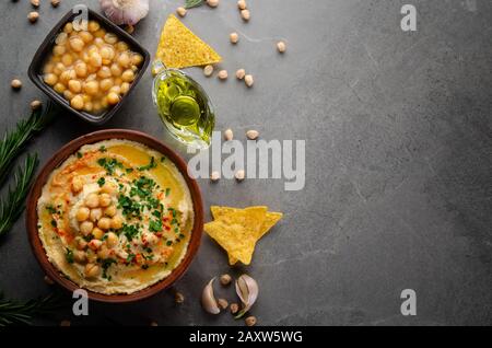 Flacher Laienblick auf Den Hummus in Tonschale mit Olivenöl, Kichererbsen und grünen Korianderblättern auf Steintisch serviert mit Ttortilla-Chips Stockfoto