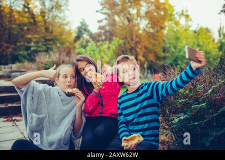 Eine Gruppe von drei Kindern, die selfie mit einem Smartphone einnehmen, während sie im Freien Pizza essen und gemeinsam Spaß haben. Kinder schaufeln mit dem Daumen nach oben. Stockfoto