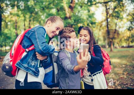 Eine Gruppe von Freunden hängt nach der Schule ab, lacht und sieht lustige Videos auf dem Smartphone, während sie durch den Park gehen. Stockfoto