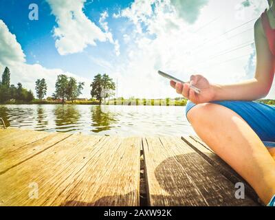 Ein Mädchen mit einem Telefon in der Hand sitzt auf der Holzoberfläche nahe der Wasseroberfläche. Von unten aufgenommen. Weitwinkelkamera für Profis. Stockfoto