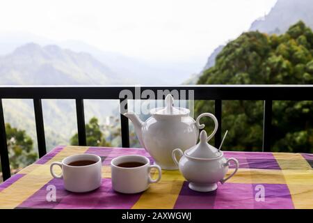 Teekanne mit Tee und Tassen auf dem Tisch mit wunderschönem Blick auf die Berge. Sri-lankanischer Tee, der am besten gebraut wird. Stockfoto