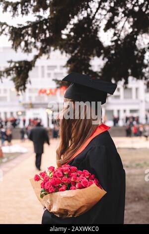 Rückansicht der Studentin der Graduierung mit einem Blumenstrauß, während sie bei der Abschlussfeier auf dem Campus steht Stockfoto