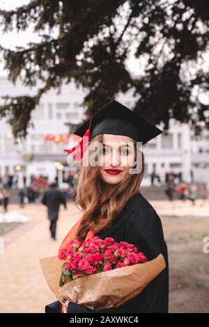 Fröhliche junge Studentin im Graduiertenkolleg, die Blumenstrauß hält, während sie bei der Abschlussfeier auf dem Campus steht Stockfoto