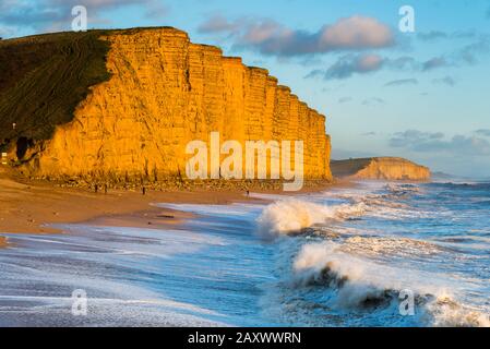 West Bay, Dorset, Großbritannien. Februar 2020. Wetter in Großbritannien. Raue Meere stürzen am East Beach in der West Bay in Dorset an Land, während die Sandsteinfelsen an einem sonnigen Nachmittag kurz vor Sonnenuntergang eine goldene Orange leuchten. Bildnachweis: Graham Hunt/Alamy Live News Stockfoto