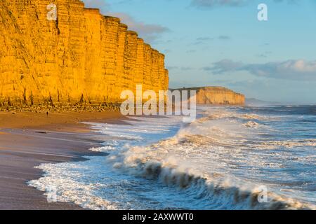 West Bay, Dorset, Großbritannien. Februar 2020. Wetter in Großbritannien. Raue Meere stürzen am East Beach in der West Bay in Dorset an Land, während die Sandsteinfelsen an einem sonnigen Nachmittag kurz vor Sonnenuntergang eine goldene Orange leuchten. Bildnachweis: Graham Hunt/Alamy Live News Stockfoto