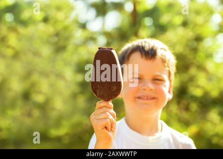 Zufriedener lächelnder Junge von 5 Jahren im Sommer draußen am heißen sonnigen Tag hält Popsicle in Schokolade auf Stock. Konzentrieren Sie sich auf Eis, Hintergrund und Kind bl Stockfoto