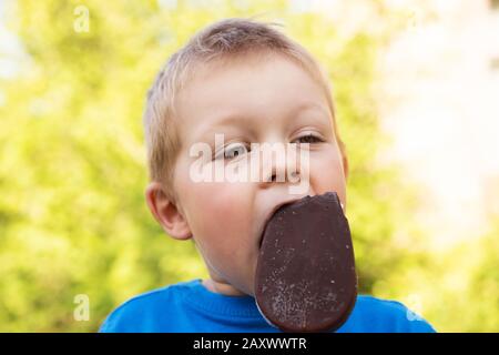 Attraktiver blonder Junge von 5 Jahren im Sommer im Freien an einem heißen sonnigen Tag, der Popsicle in Schokolade auf einem Stock isst. Nahaufnahme des Porträts auf verschwommener Backgrou Stockfoto