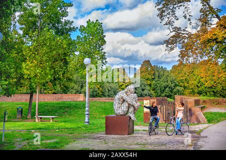 Junge Leute auf dem Fahrrad machen am sonnigen Herbsttag ein Foto in der Nähe zeitgenössischer Skulpturen im öffentlichen Park. Lucca, Toskana, Italien Stockfoto