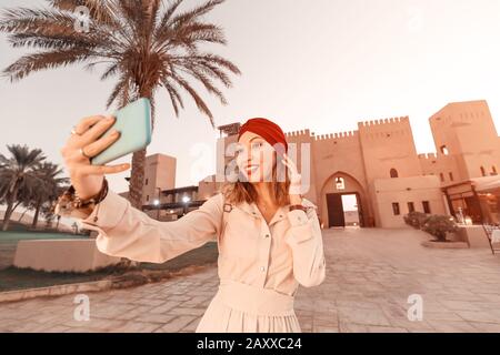 Fröhliche Frau Reisende, die Kleid und Turban trägt, fotografiert selfie auf ihrem Smartphone einer alten arabischen Stadt oder eines Dorfes mitten in der Wüste. Konce Stockfoto