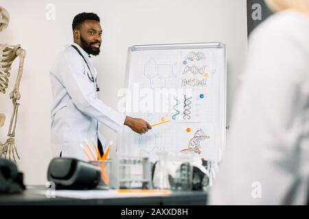 Portrait des jungen männlichen afrikanischen Arztes oder Biochemikers, der an Bord einige biochemische Formeln für seine Kollegen im Konferenzraum zeigt. Medizin Stockfoto