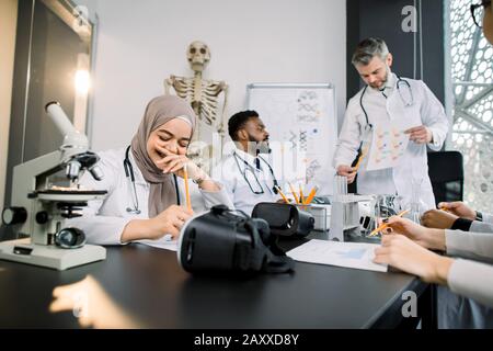 Ansehnlicher männlicher Professor mit multiethnischen Medizinstudenten oder Wissenschaftlern, die Labormäntel im Klassenzimmer tragen. Das junge, hübsche muslimische Mädchen ist glücklich und lächelnd Stockfoto