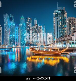 Bunte Lichter der Nachtstadt im Dubai Marina Viertel. Stilisiertes arabisches Schiff Abra Dhow mit Lichtern im Vordergrund. Stockfoto