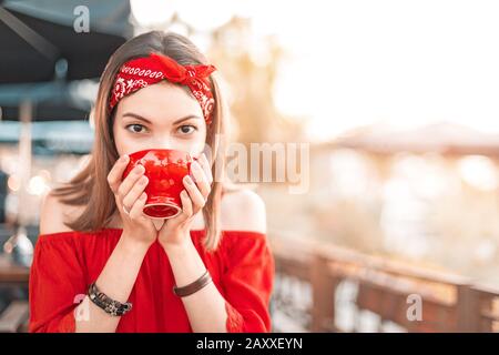 Fröhliche asiatin in Rot, die in einem Terrassencafé heißen Tee trinkt Stockfoto