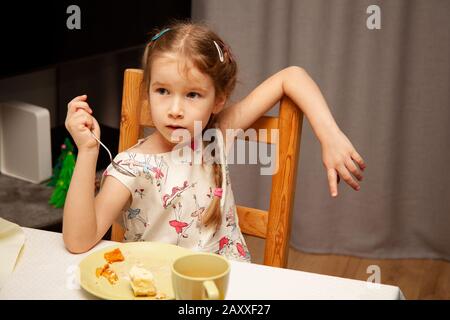 Kleines junges Mädchen, das ihr Essen isst und eine Gabel in der Hand hält, die auf einem Stuhl in einer schlampigen, unachtsamen entspannten Haltung/Art sitzt. Arm auf der Rückseite des Stuhls Stockfoto