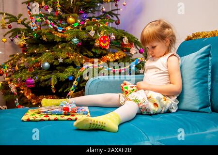 Kleines junges Mädchen, das am bunten weihnachtsbaum sitzt und ihre Geschenke auspacken und auspacken kann. Halten Sie ein Paket in den Händen, einige Kisten davor Stockfoto