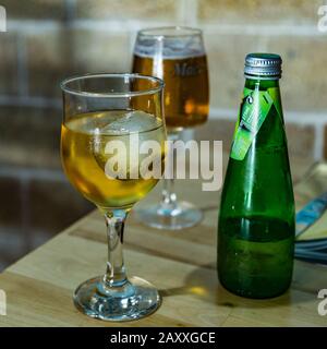 Auf dem Esstisch befindet sich ein Glas kalter, funkelnder apfelsaft mit Eiswürfel und grüner Flasche. Kleines schaumiges Bier auf unscharfem Hintergrund. Selektiv Stockfoto