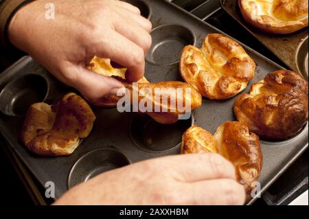 Schließen Sie sich den Händen eines männlichen Küchenchefs an, und entfernen Sie gekochte individuelle goldene Yorkshire Puddings aus einem Backblech, das frisch aus dem Ofen stammt Stockfoto