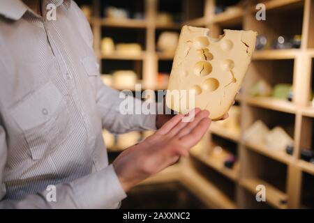 Hansome Mann hält große Scheibe Käse maasdam in der Hand. Käse mit großen Löchern. Hintergrund der Regale mit Käse Stockfoto