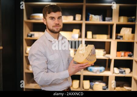 Hansome Mann hält große Scheibe Käse maasdam in der Hand. Käse mit großen Löchern. Hintergrund der Regale mit Käse Stockfoto