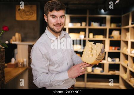 Hansome Mann hält große Scheibe Käse maasdam in der Hand. Käse mit großen Löchern. Hintergrund der Regale mit Käse Stockfoto