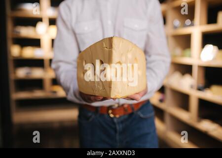 Hansome Mann hält große Scheibe Käse maasdam in der Hand. Käse mit großen Löchern. Hintergrund der Regale mit Käse Stockfoto