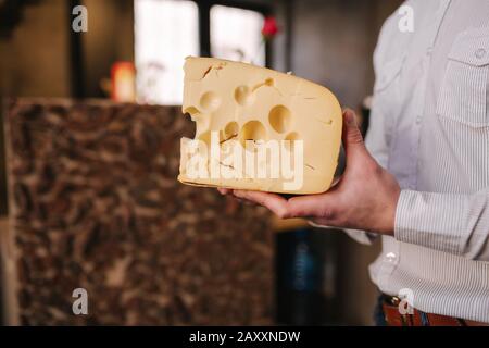 Hansome Mann hält große Scheibe Käse maasdam in der Hand. Käse mit großen Löchern. Hintergrund der Regale mit Käse Stockfoto