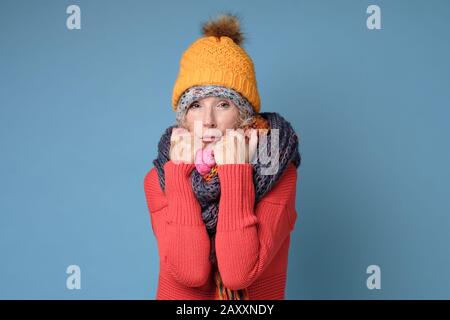 Gefrorene Seniorin in Hüten und Narben isoliert an blauer Wand. Kaltwetterkonzept Stockfoto