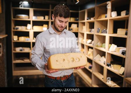 Hansome Mann hält große Scheibe Käse maasdam in der Hand. Käse mit großen Löchern. Hintergrund der Regale mit Käse Stockfoto