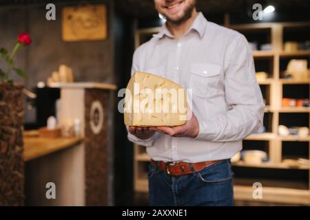 Hansome Mann hält große Scheibe Käse maasdam in der Hand. Käse mit großen Löchern. Hintergrund der Regale mit Käse Stockfoto