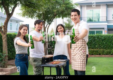 Asiatischer Mann, der grillt und Wurst für eine Gruppe von Freunden kocht, um im Garten zu Hause Party zu essen. Gruppe von Freunden, die Gartengrill la im Freien haben Stockfoto