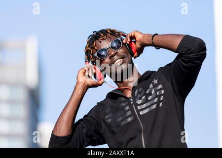 Vorderansicht des schwarzen Mannes mit legerer Kleidung und Sonnenbrille, die auf der Straße stehen, während sie mit Kopfhörern Musik an sonnigen Tagen hören Stockfoto