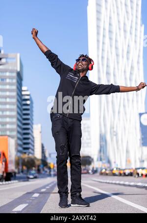 Vorderansicht des schwarzen Mannes mit legerer Kleidung und Sonnenbrille, die auf der Straße stehen, während sie mit Kopfhörern Musik an sonnigen Tagen hören Stockfoto