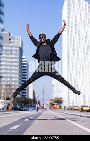 Vorderansicht junger schwarzer Mann mit legeren Kleidungsstücken, die im städtischen Hintergrund springen. Lifestyle-Konzept. Tausendjähriger afrikanischer Kerl trägt Sonnenbrille im Freien Stockfoto