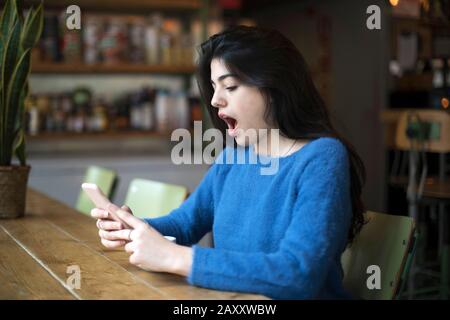 Seitenansicht eines legeren Mädchens, das Smartphone beobachtet und beim Sitzen in der Cafeteria mit Neuigkeiten überrascht aussieht Stockfoto