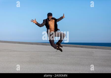 Vorderansicht junger schwarzer Mann mit legeren Kleidungsstücken, die im städtischen Hintergrund springen. Lifestyle-Konzept. Tausendjähriger afrikanischer Kerl trägt Sonnenbrille im Freien Stockfoto