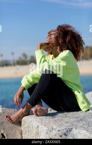 Seitenansicht der schönen lockigen afro-Frau, die auf Wellenbrecher sitzt, lacht, während sie unter den Kinnaugen, die im Freien geschlossen sind, die Hand ruht Stockfoto