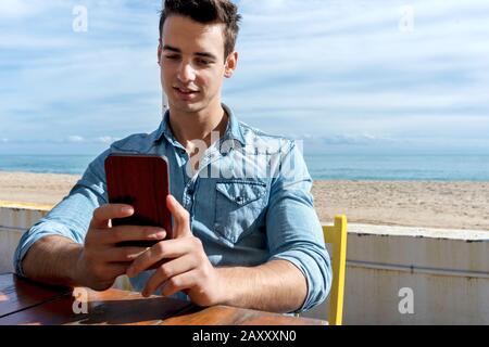 Vorderansicht des jungen Mannes, der an einem sonnigen Tag im Strandclub sitzt und dabei ein Handy benutzt Stockfoto