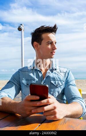 Vorderansicht des jungen Mannes, der an einem sonnigen Tag im Strandclub sitzt und dabei ein Handy benutzt Stockfoto