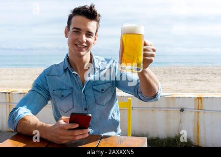 Vorderansicht des jungen Mannes, der an einem sonnigen Tag im Strandclub sitzt und dabei ein Handy benutzt Stockfoto